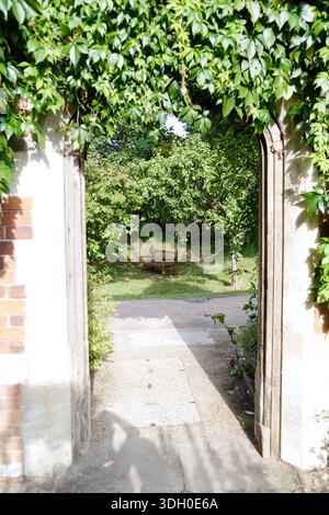 A tranquil garden scene with a wooden bench viewed through a stone archway draped in lush green ivy. Concept of peace and contemplation. Stock Photo