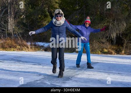 Two senior women sliding and having fun on a frozen lake in winter, enjoying outdoor activity and friendship in a scenic snowy landscape Stock Photo
