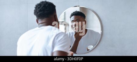 Man Applies Skincare Product and Smiles While Looking in Mirror at Home Bathroom Stock Photo