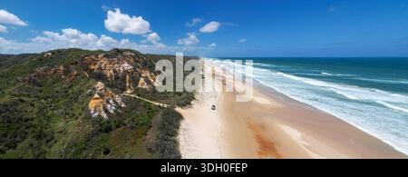 Aerial view of a lone 4wd car driving on a 75 Mile Beach where the turquoise ocean meets the shore, and the cliffs rise with green vegetation surround Stock Photo