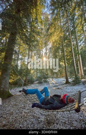 Woman relaxing in a hammock in a quiet winter forest, enjoying the silence as warm sun rays filter through the trees, creating a peaceful and mindful Stock Photo