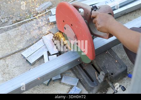 Focused male construction worker cutting steel beam with red chop saw at industrial site using metal cutter with care and precision to spark bright fi Stock Photo