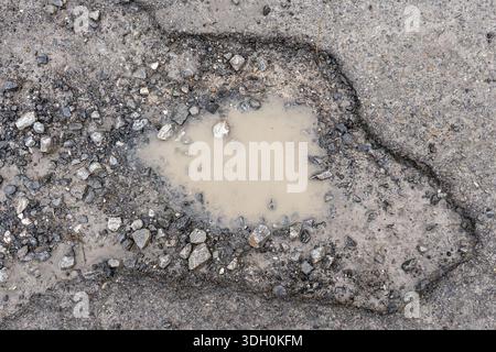 Muddy puddle filling a pothole on a damaged asphalt road surrounded by cracked pavement and loose gravel. Concept of urban infrastructure deterioratio Stock Photo