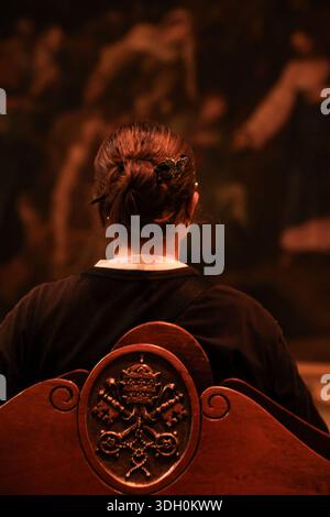 Young woman sitting on a wooden bench with the Vatican coat of arms, contemplating classical paintings in the Pinacoteca Vaticana, Vatican Museums. Stock Photo