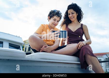 Mother and son using a smartphone together outdoors on a boat, sharing a relaxed moment. Concept of family connection, technology in daily life, commu Stock Photo