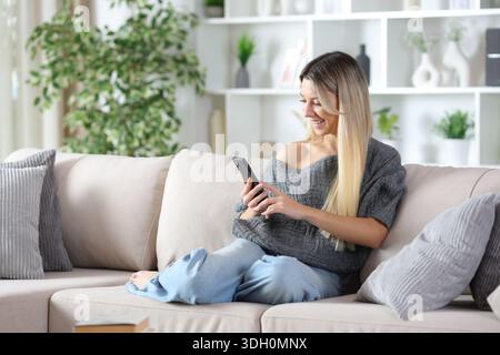 Happy woman enjoying using smart phone sitting on a couch at home Stock Photo
