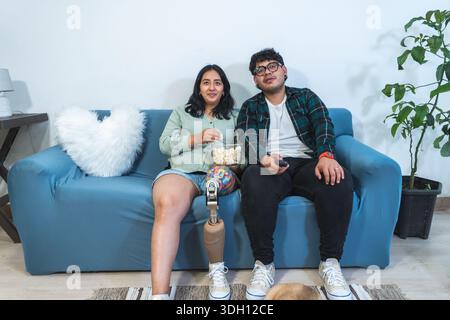 Happy hispanic couple enjoying a movie night at home, eating popcorn and watching television Stock Photo