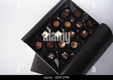 top-down view of an open black gift box filled with assorted artisan chocolates in glossy black compartments on a minimalist white backdrop, evoking e Stock Photo