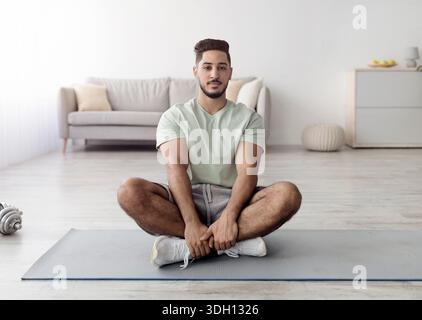 Balanced young Arab guy sitting cross legged on sports mat, meditating or practicing yoga at home, full length Stock Photo