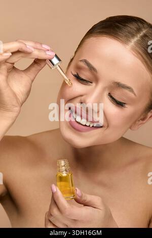 A woman is using a dropper bottle to apply facial oil to her skin. She is smiling and showing a positive expression. The setting has a neutral backgro Stock Photo