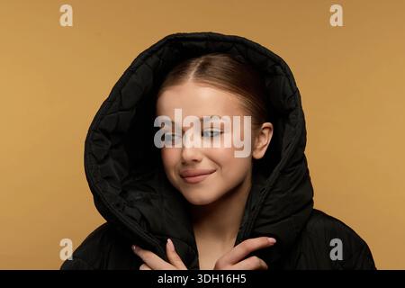A young woman wears a black hoodie with a large hood and smiles while posing against a warm yellow background in a studio setting. Her hair is styled Stock Photo