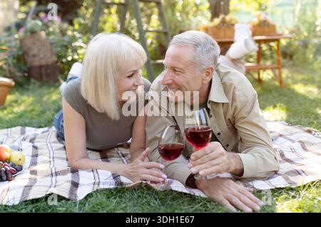 Loving married senior couple drinking wine outside, lying on blanket while having picnic in their garden Stock Photo