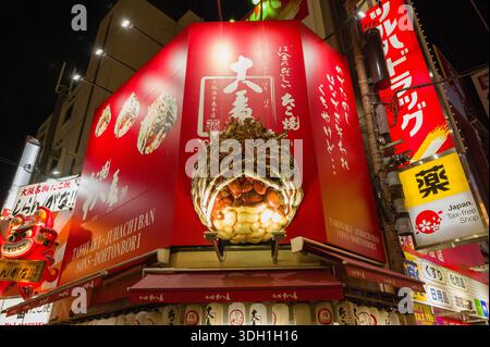Osaka, Japan - November 5, 2023: Large takoyaki sculpture and illuminated red signage above the entrance of Takoyaki Juhachiban restaurant in the Dotonbori district at night, with Japanese writing, commercial logos, and neighboring storefronts visible. Stock Photo