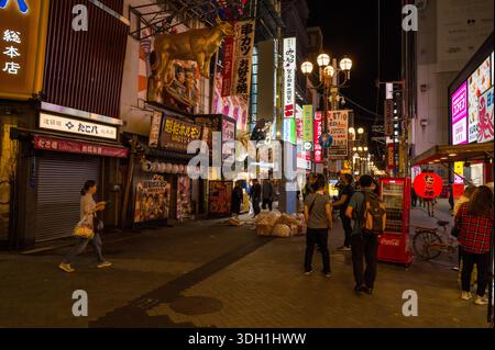 Osaka, Japan - November 5, 2023: People walk along a commercial street at night in the Dotonbori district with illuminated shop signage, restaurant storefronts, a large golden cow sign, vending machines, and garbage bags visible. Stock Photo