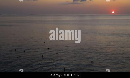 Aerial view of surfers dotting the tranquil, twilight sea under a fiery sunset sky, Bali, Bali, Indonesia. Stock Photo