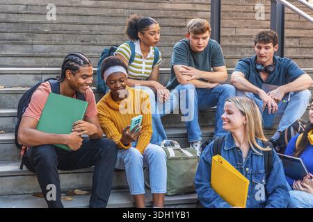 Diverse group of university students are relaxing and interacting on campus stairs, sharing moments and looking at a smartphone, embodying youth, frie Stock Photo