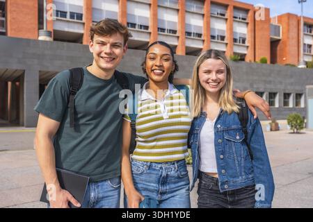 Three young multiethnic students with backpacks posing and smiling at the camera, standing in front of a modern brick university building on a sunny d Stock Photo
