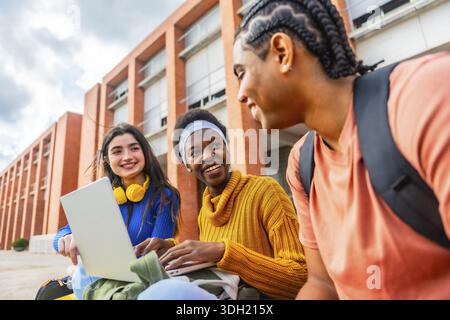 Diverse group of university students collaborating and smiling around a laptop outdoors on campus, sharing ideas, studying and enjoying casual teamwor Stock Photo