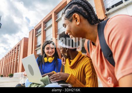 Three diverse students are working together on a laptop computer outside a university building, showcasing teamwork, learning, and connection amongst Stock Photo