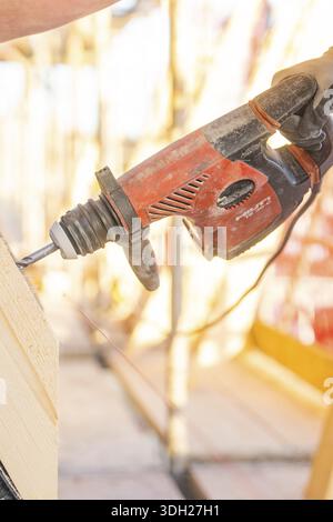 A construction worker operates a red drill on a construction site. Sunlight illuminates the scene, carpentry construction site, roof extension, renova Stock Photo