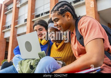 Group of happy young diverse students collaborating on a project, smiling and looking at a laptop computer outdoors at a university, highlighting team Stock Photo