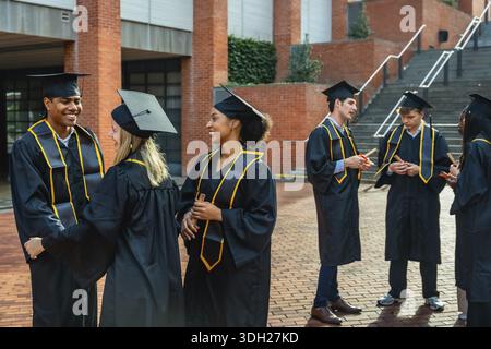 Happy diverse students wearing graduation caps and gowns, holding diplomas, and conversing outdoors on a university campus after their commencement ce Stock Photo