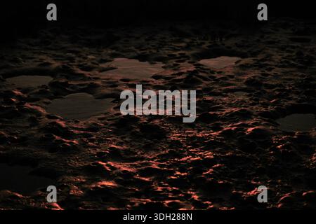 Sunset light reflecting on wet mudflat surface, organic earth textures and glowing highlights forming an abstract natural background, dark tones Stock Photo