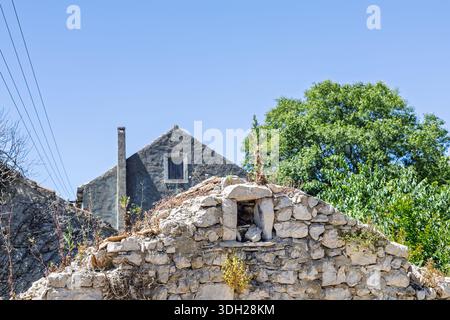 A weathered stone house sits behind a crumbling stone wall, beneath a clear blue sky. The scene captures rural charm, aging architecture, and hardy ve Stock Photo