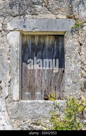 A rustic stone wall frames a weathered wooden window, with ivy and plants growing along the top and sides. Two glass cups rest on a sunlit ledge, evok Stock Photo