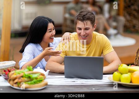 Young multinational couple using laptop near camper van outdoors, chatting with friends online while having breakfast Stock Photo