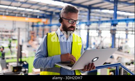 Quality manager with headset and laptop standing in industrial factory. Stock Photo