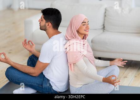 Yoga for pregnancy. Happy muslim spouses meditating together at home, sitting back to back and practicing lotus pose Stock Photo