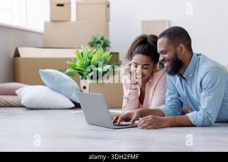 Smiling young african american woman and man looking at laptop with new design for room, lying on floor with boxes Stock Photo