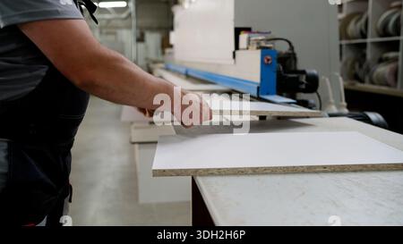 Close-up of a worker's hands taking processed wood panels from an edge bander machine in a busy furniture manufacturing workshop Stock Photo