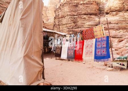 Brightly colored fabrics are displayed for sale on a line in a dry desert area surrounded by large rock formations. Tents provide shade for visitors a Stock Photo