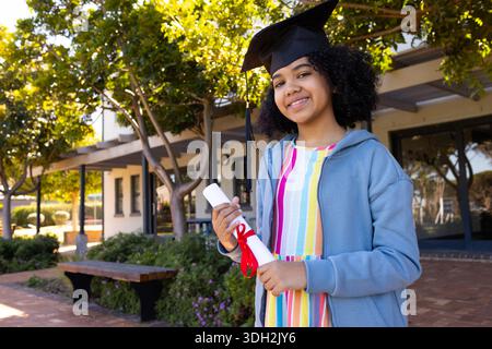 African teen female student standing on campus brick walkway wearing mortarboard holding diploma Stock Photo
