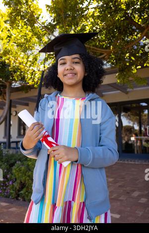 African American teen female in hoodie standing on campus holding cap and diploma with red ribbon Stock Photo