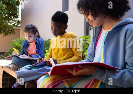 Diverse children sitting on low stone bench in courtyard with backpacks, marking and writing notes Stock Photo