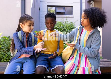 Diverse children sitting on wooden bench in courtyard playing hand game, wearing colorful backpacks Stock Photo