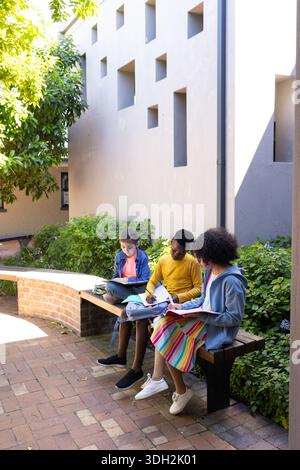Diverse teenage students studying together on wooden bench in courtyard with notebooks Stock Photo