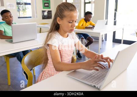 Classmates typing at white desks using silver laptops in classroom with girl in pink gingham dress Stock Photo