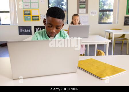 Diverse students working on laptops in class with silver laptop, notebook, green shirt, pink dress Stock Photo