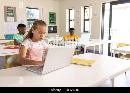 Diverse classmates sitting at white tables using silver laptops, female youth with yellow notebook Stock Photo