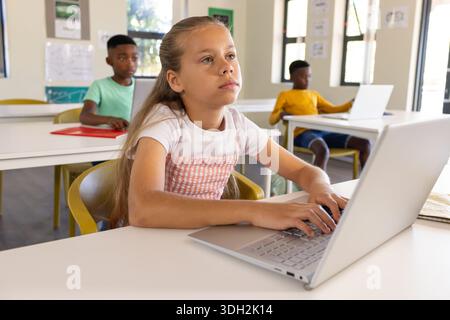 Diverse youth students sitting in classroom, female youth in pink dress typing on silver laptop Stock Photo