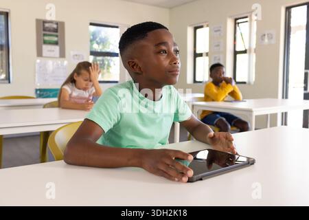 Diverse youth classmates learning at white desks under windows, boy in light green tee using tablet Stock Photo