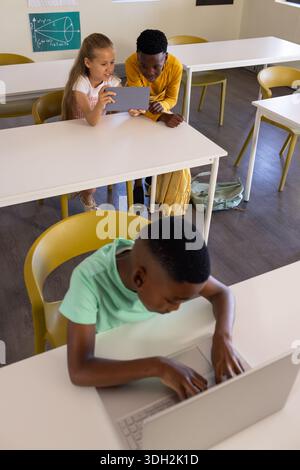 Diverse classmates sharing tablet while boy in mint T-shirt typing on silver laptop in classroom Stock Photo