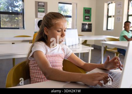 Diverse classmates including female 10-12yr child wearing pink pinafore using laptop in classroom Stock Photo