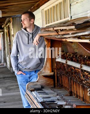 Man poses besides a broken upright piano.  He is looking away lost in thought.  Front is missing from piano and insides exposed to the elements. Stock Photo