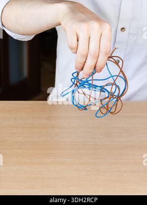 Close-up of a hand holding knotted electrical cables in blue and brown colors over a light wooden surface, symbolizing wiring confusion. Stock Photo