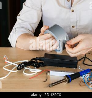 Person's hands actively tearing a piece of gray duct tape in a workspace filled with electronic wires, soldering iron, and adapters on a wooden desk. Stock Photo